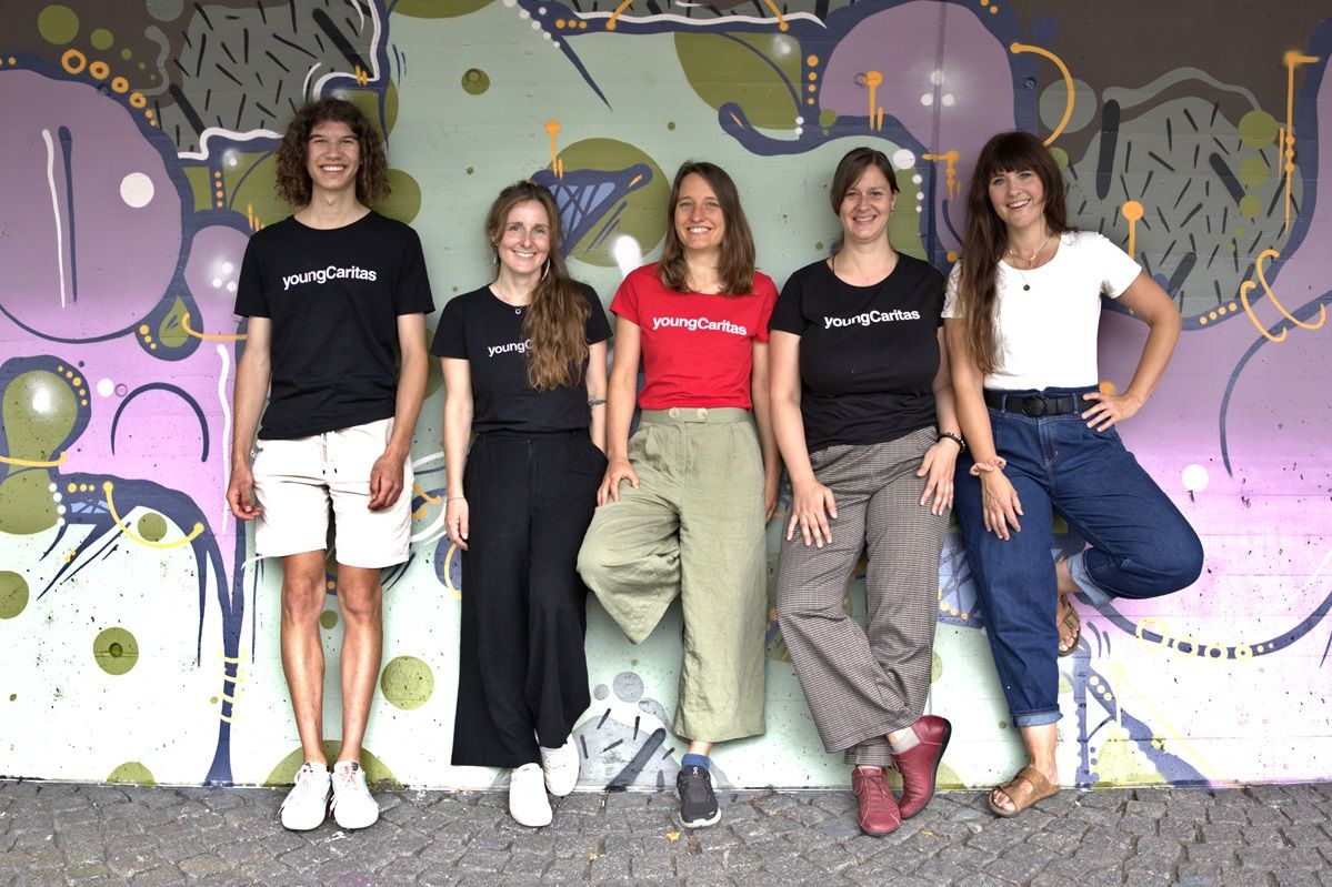 Fünf junge Erwachsene posieren vor einer bunten Wand mit dem Logo von youngCaritas im Hintergrund.