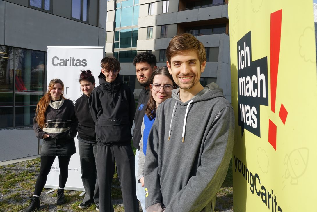 Eine Gruppe junger Menschen steht vor einem Caritas-Schild, lächelnd und bereit für soziale Aktivitäten.