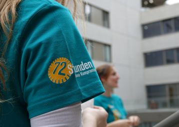 Nahaufnahme eines T-Shirts mit 72 Stunden ohne Kompromiss-Logo. Im Hintergrund sind weitere Personen und Gebäude sichtbar.