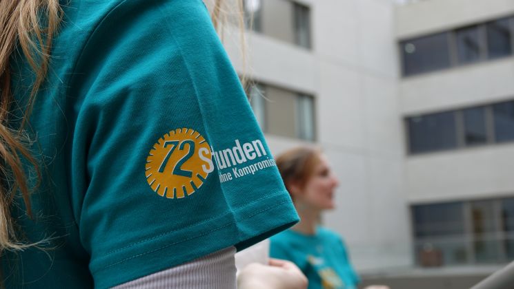 Nahaufnahme eines T-Shirts mit 72 Stunden ohne Kompromiss-Logo. Im Hintergrund sind weitere Personen und Gebäude sichtbar.