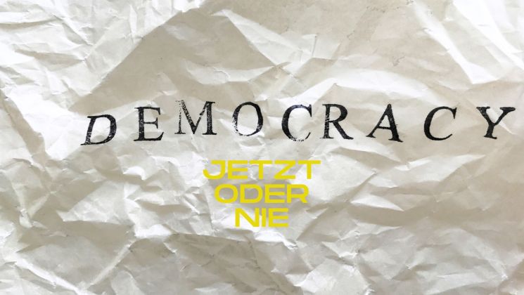 Ein zerknittertes Blatt mit dem Wort 'DEMOCRACY' und der Aufschrift 'JETZT ODER NIE' in gelber Farbe.