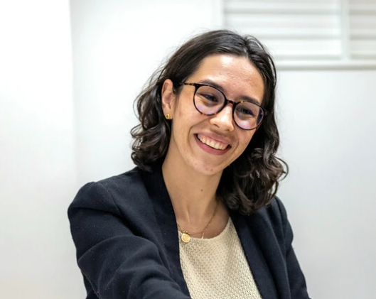 Eine Frau mit lockigem Haar und Brille lächelt freundlich in einem modernen Büro. Sie trägt einen schwarzen Blazer und hat einen warmen, einladenden Ausdruck.
