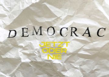 Ein zerknittertes Blatt mit dem Wort 'DEMOCRACY' und der Aufschrift 'JETZT ODER NIE' in gelber Farbe.
