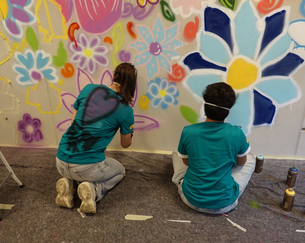 Zwei Personen in blauen T-Shirts malen bunte Blumen und Muster an eine Wand. Spraydosen liegen neben ihnen.