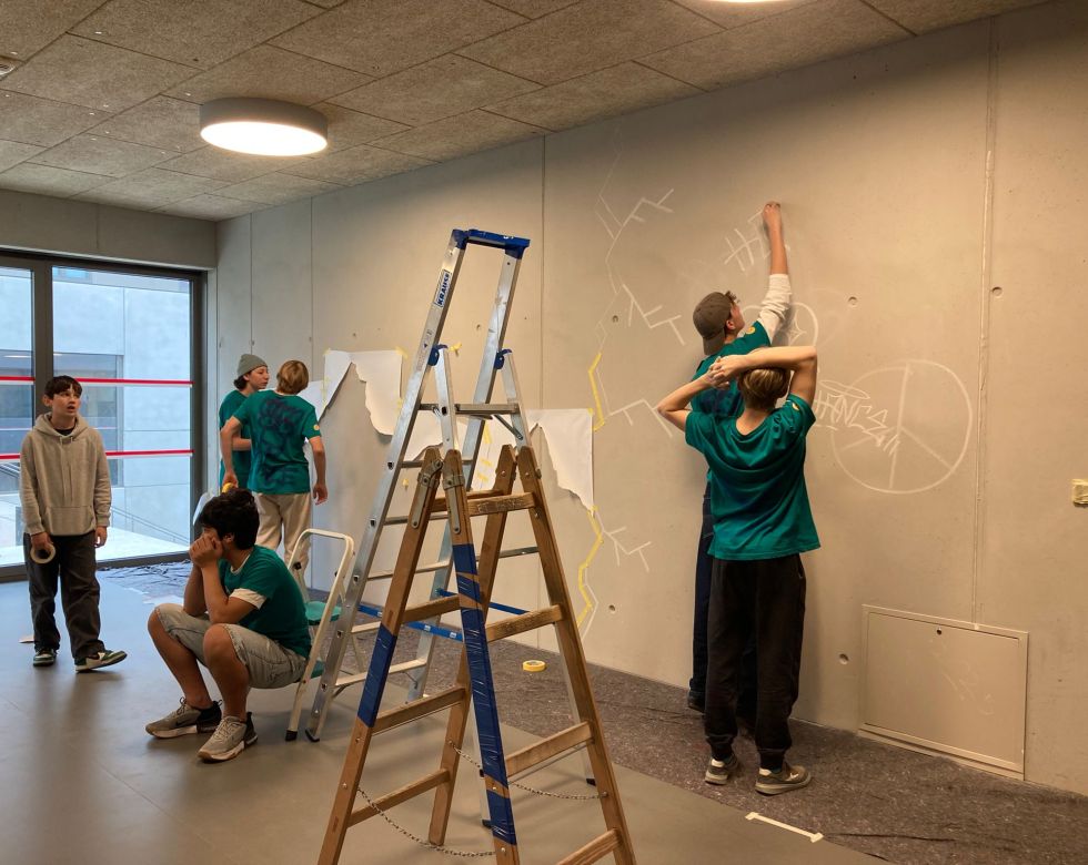 Eine Gruppe von Schülern gestaltet eine Wand mit Skizzen, während eine Leiter im Vordergrund steht.