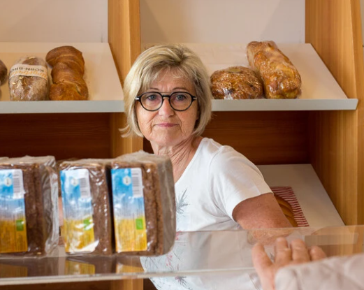 Eine Frau mit Brille steht an einer Theke in einer Bäckerei, mit frisch gebackenem Brot im Hintergrund.