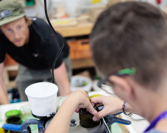 Zwei Personen arbeiten in einer Werkstatt mit Glasgefäßen, während eine Person eine Maschine bedient und die andere sie beobachtet.