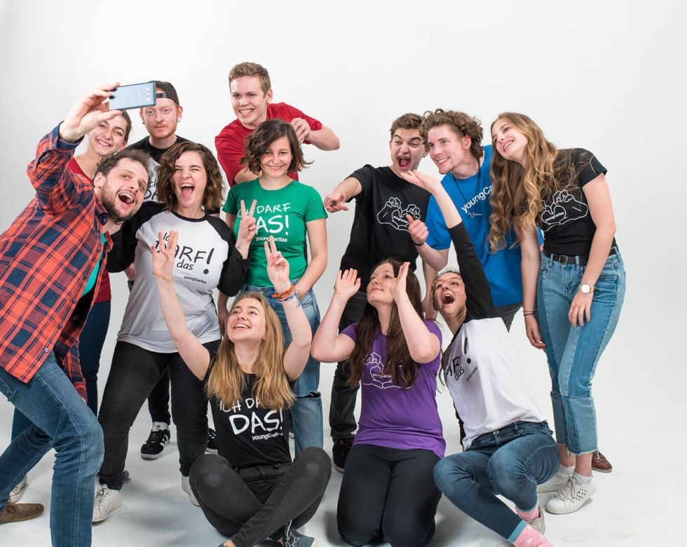 Eine Gruppe von jungen Menschen macht ein fröhliches Selfie in einem Studio, alle tragen T-Shirts mit verschiedenen Aufdrucken.