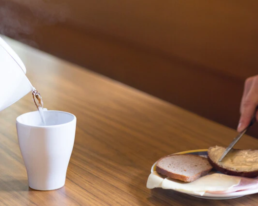 Eine Hand schmiert Butter auf ein Stück Brot, während ein Wasserkocher heißes Wasser in eine Tasse gießt.