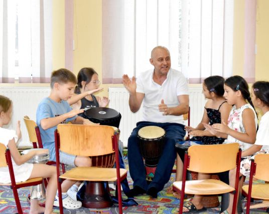 Ein Lehrer zeigt einer Gruppe von Kindern, wie man verschiedene Trommeln spielt. Die Kinder sind aufmerksam und haben Spaß am Musizieren.