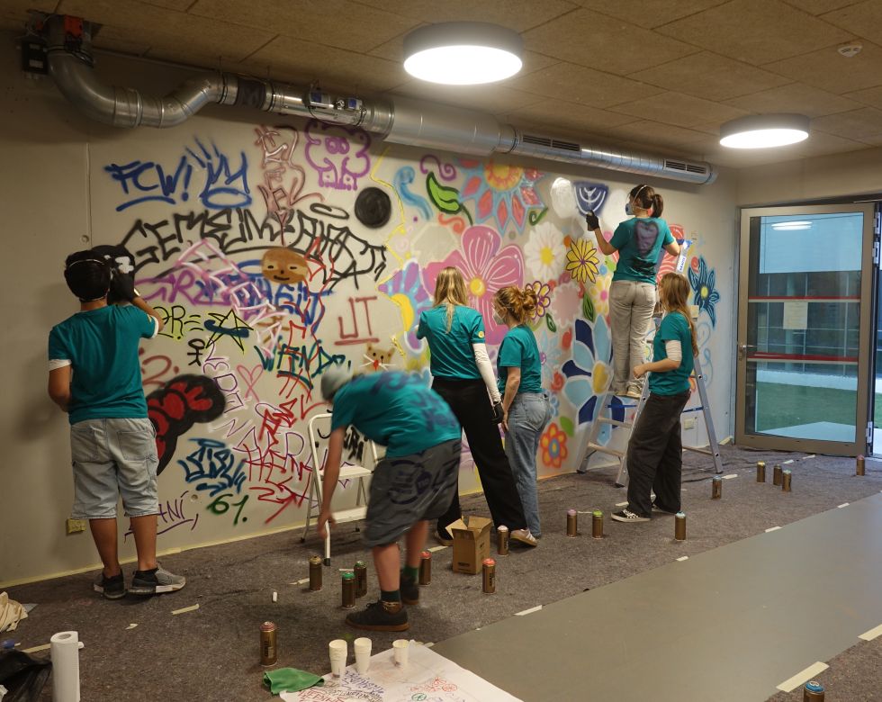 Eine Gruppe junger Menschen malt farbenfrohes Graffiti an einer Wand, während sie mit verschiedenen Farben und Sprays arbeiten.