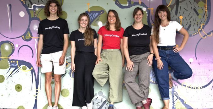 Fünf junge Erwachsene posieren vor einer bunten Wand mit dem Logo von youngCaritas im Hintergrund.