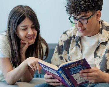 Ein Junge und ein Mädchen sitzen zusammen und lesen ein Buch. Der Junge trägt eine Brille und die beiden schauen interessiert ins Buch.