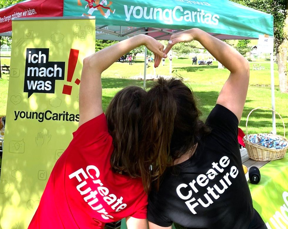 Zwei junge Frauen formen ein Herz mit ihren Händen, während sie Shirts mit den Aufschriften „Create Future“ tragen. Im Hintergrund ein Stand von youngCaritas.
