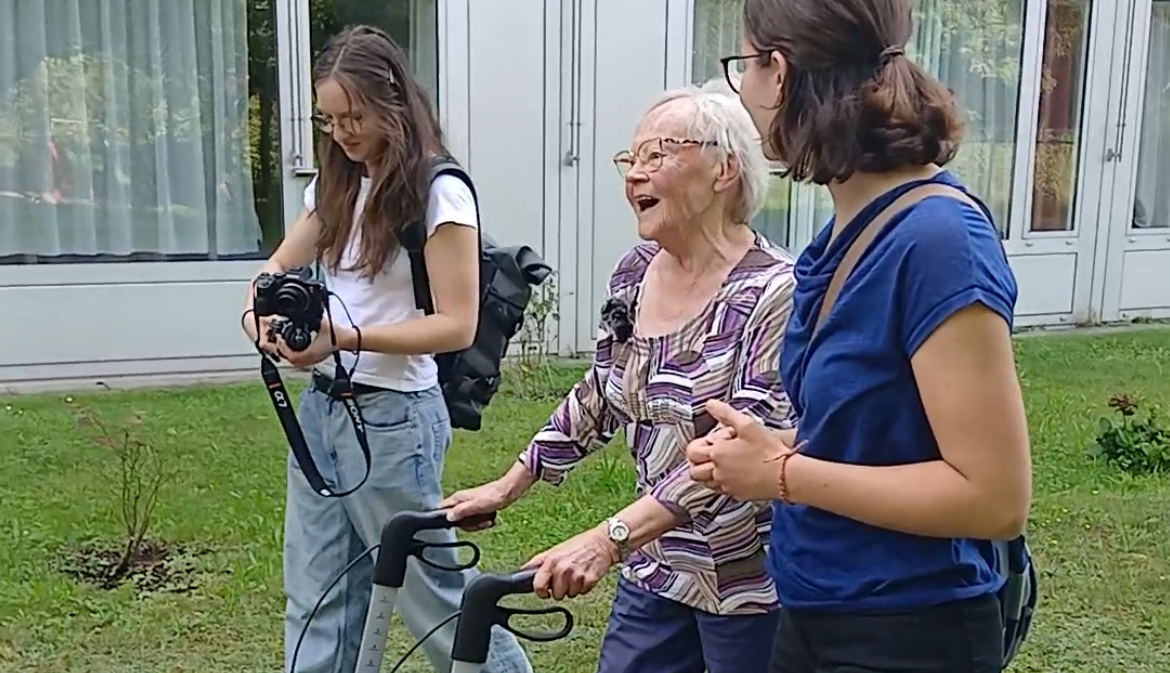 Eine ältere Frau benutzt einen Rollator im Freien, um mit zwei jungen Frauen zu interagieren. Sie lachen und genießen die Zeit.
